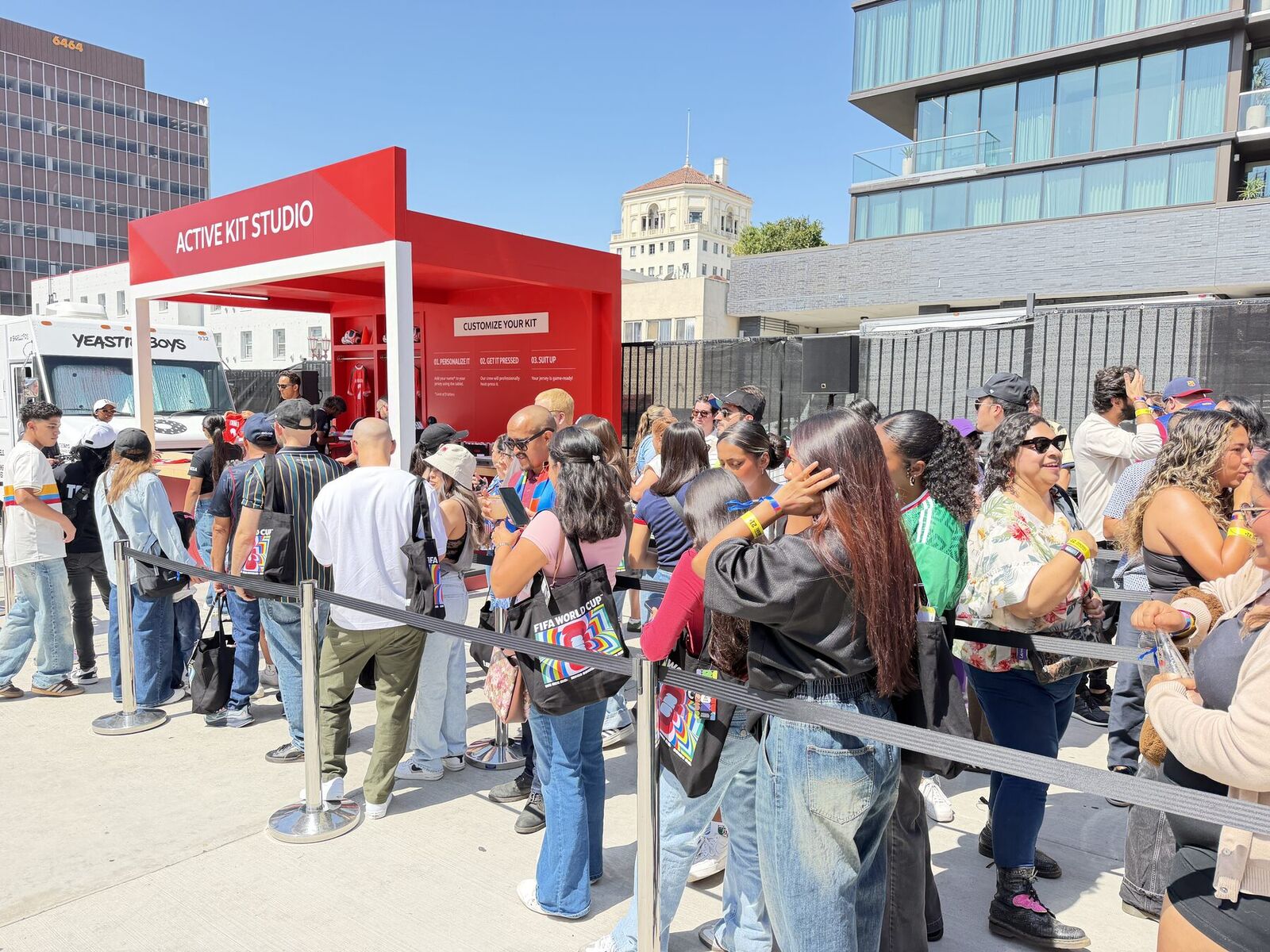 A long line of guests waiting behind stanchions at a red outdoor jersey personalization studio on a sunny plaza